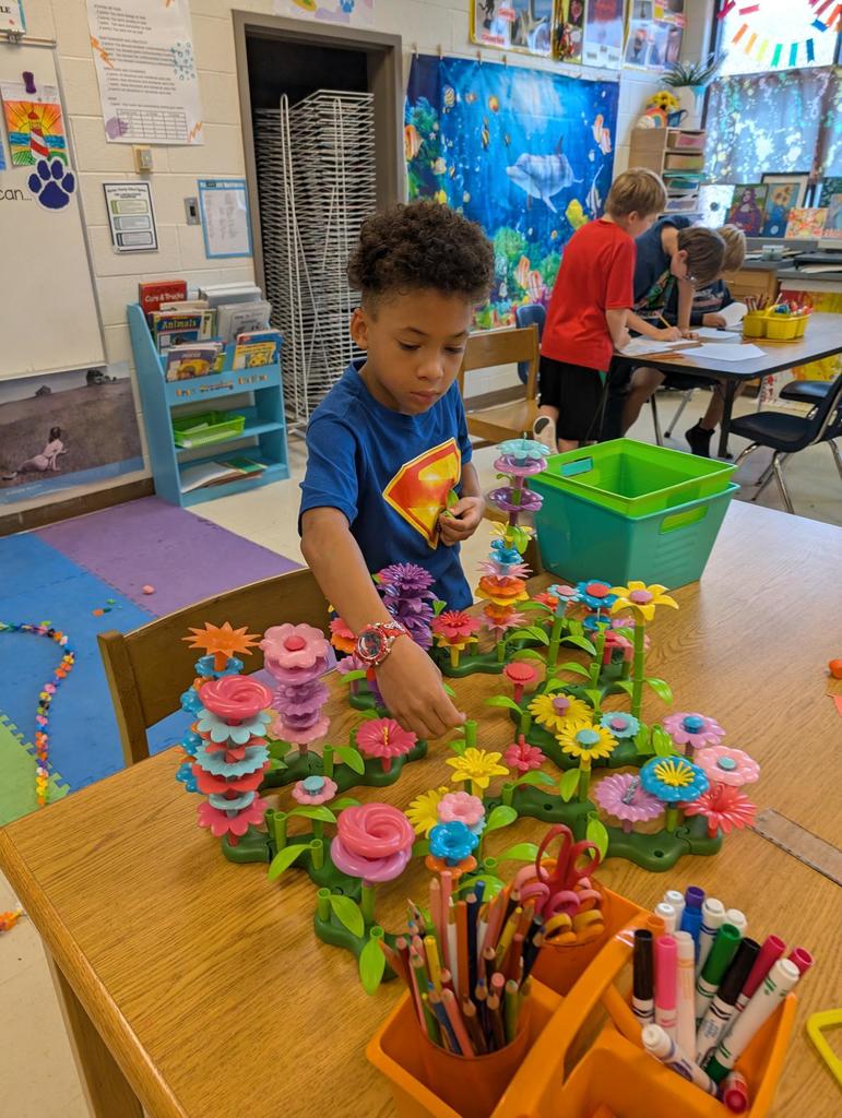 There is a boy in a Superman shirt looking at some flowers made from Legos.