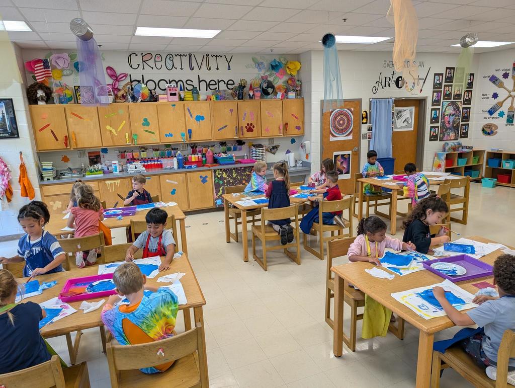 There are five tables with boys and girls at them drawing a picture in art class.  They are using markers and stencils.