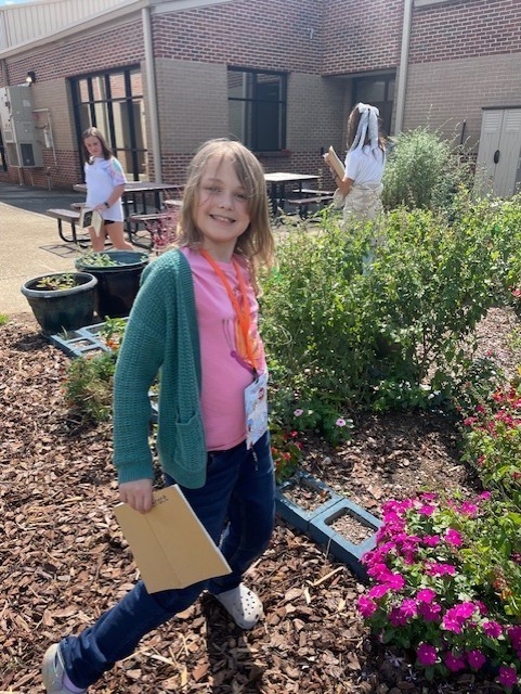 A girl in a pink shirt and green jacket is smiling.  She is holding a folder in her hand.  There are two girls in the background working in the garden.