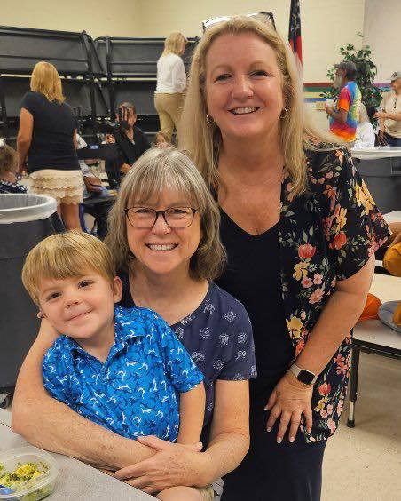 2 grandmothers and a little kid  posing for the camera at grandparents day lunch. There is a tray of food at the table in front on them..