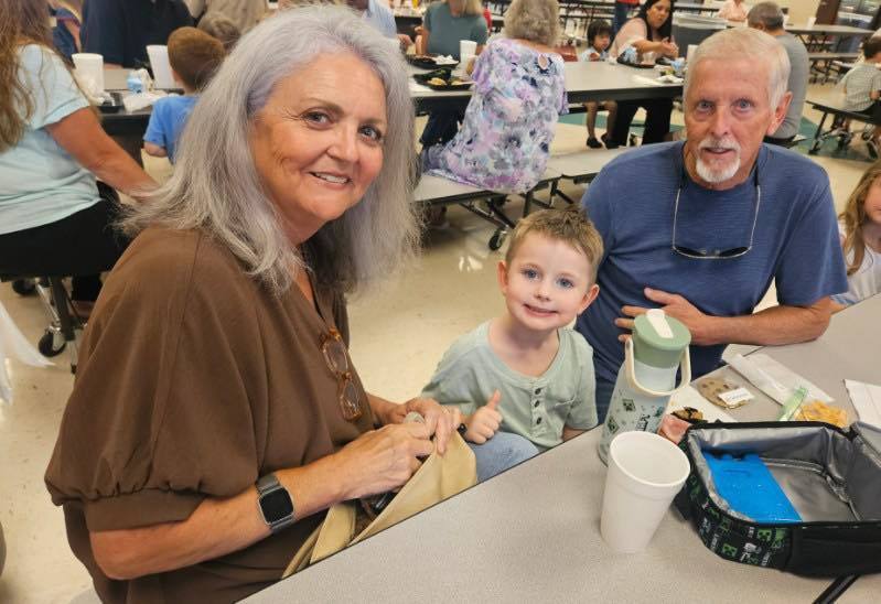 A grandmother and a grandfather and a little kid  posing for the camera at grandparents day lunch. There is a tray of food at the table in front on them..