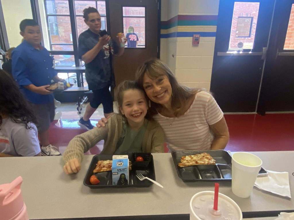A grandmother and a little kid  posing for the camera at grandparents day lunch. There is a tray of food at the table in front on them..