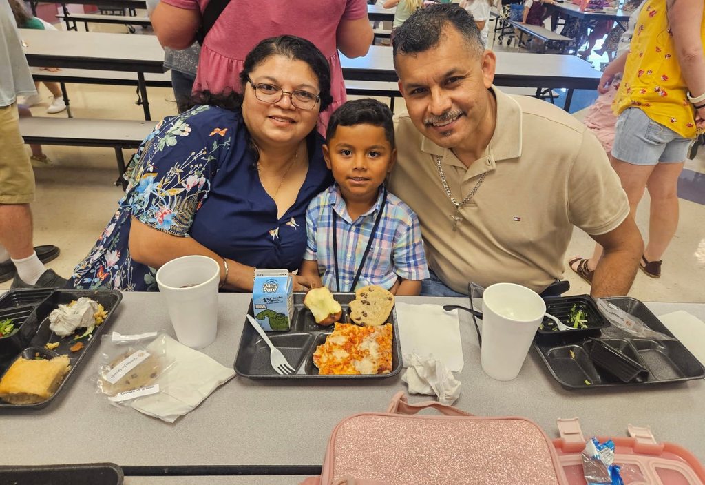 A grand father and a grandmother and a little kid  posing for the camera at grandparents day lunch. There is a tray of food at the table in front on them..