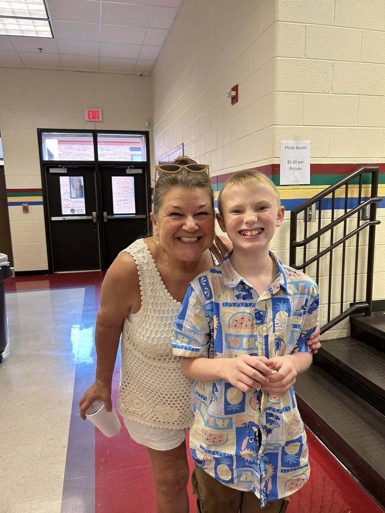 A grandmother and a little kid  posing for the camera at grandparents day lunch. There is a tray of food at the table in front on them..