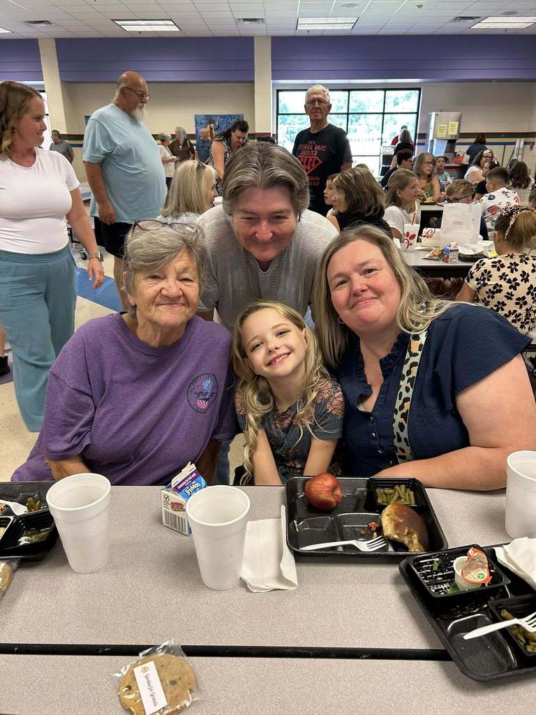 3  grandmothers and a little kid  posing for the camera at grandparents day lunch. There is a tray of food at the table in front on them..