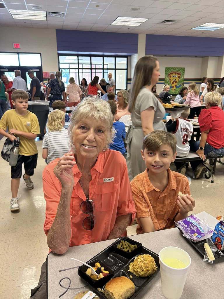 A grandmother and a little kid  posing for the camera at grandparents day lunch. There is a tray of food at the table in front on them..