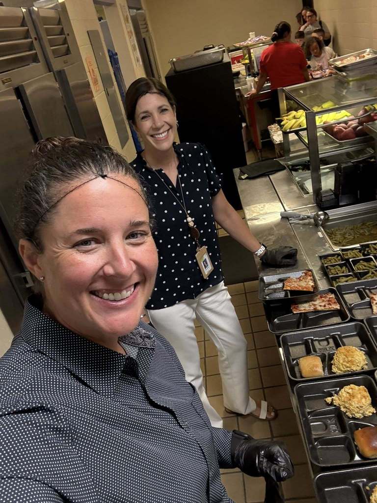 2 women wearing hairnets serving food on black trays. 