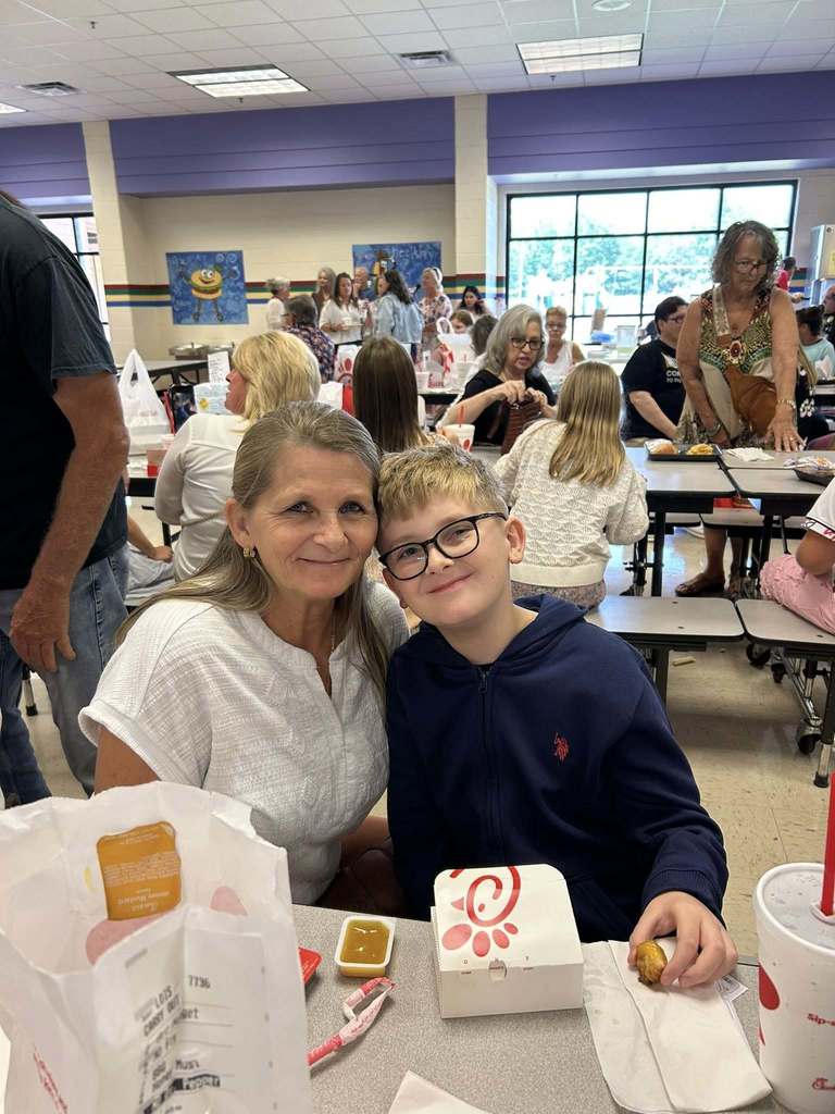 A grandmother and a little kid  posing for the camera at grandparents day lunch. There is a tray of food at the table in front on them..