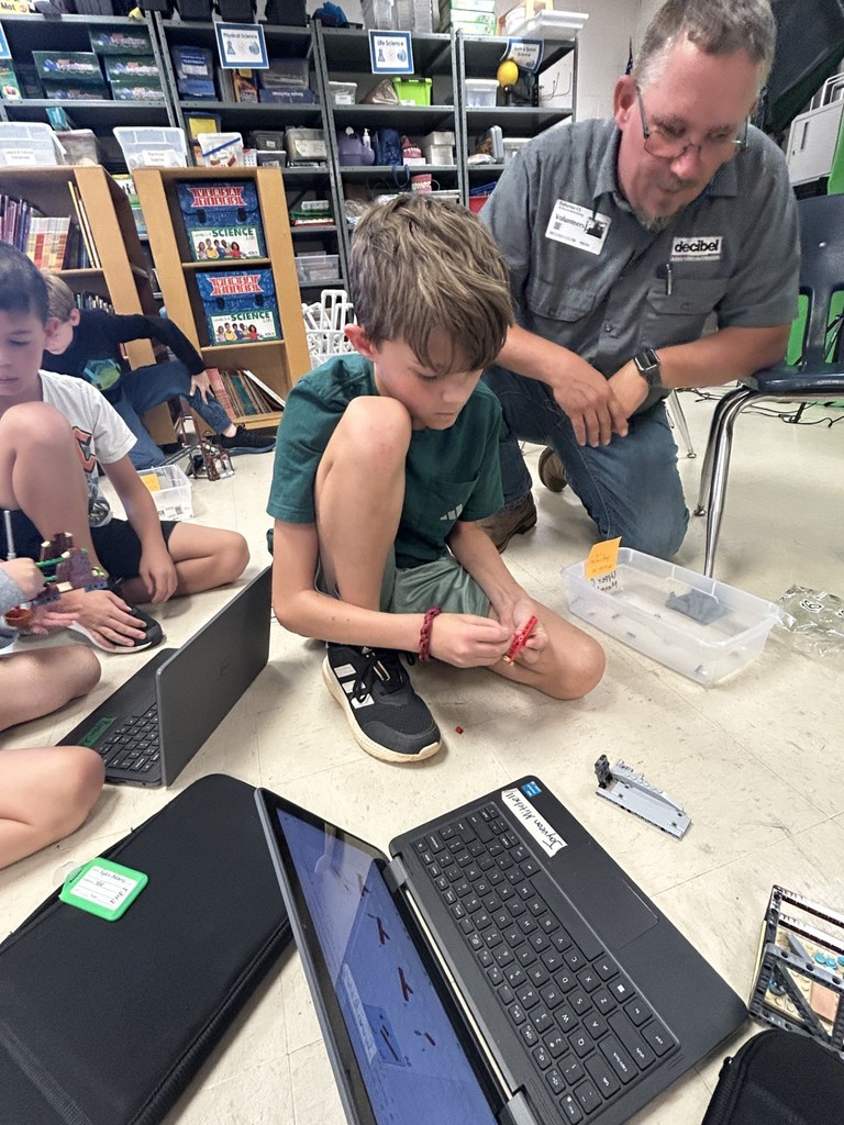 There is a parent (a man) bent down talking to a student (a boy) on the floor.  The student has his laptop open and a piece of red plastic in his hand.