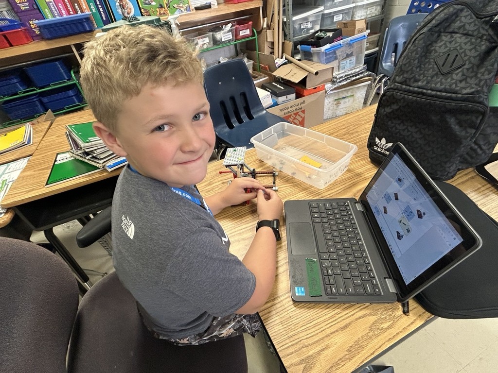 There is a boy sitting at a desk with his laptop open.  The student is smiling for the camera and there is  an airplane robot in his hands.