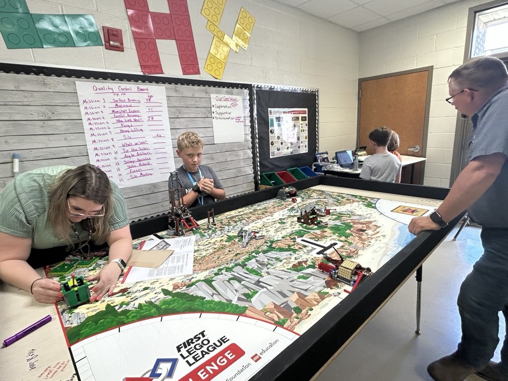There is a lady teacher and a male parent watching a student (a boy) work on a robot.  He is standing behind a  Lego tables  with landforms on it.  The lady teacher is trying to turn on one robot.