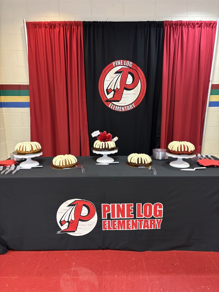 table with a black table cloth with “Pine Log Elementary” logo on it. on the table are 5 cakes