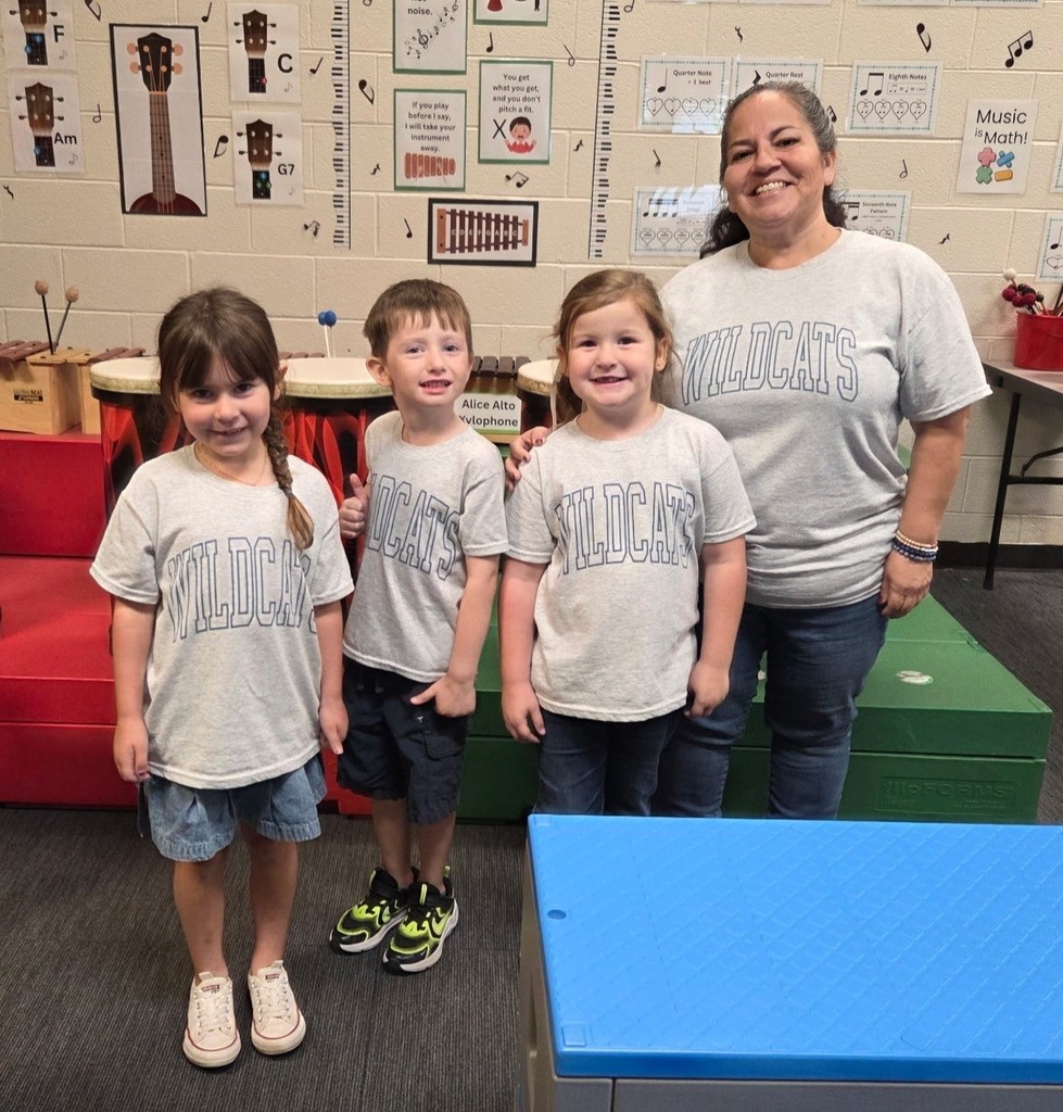The teacher, Mrs. Pettiti, is standing with two girls and a boy in a classroom.  They are all wearing a grey shirt that says "Wildcats" in blue.