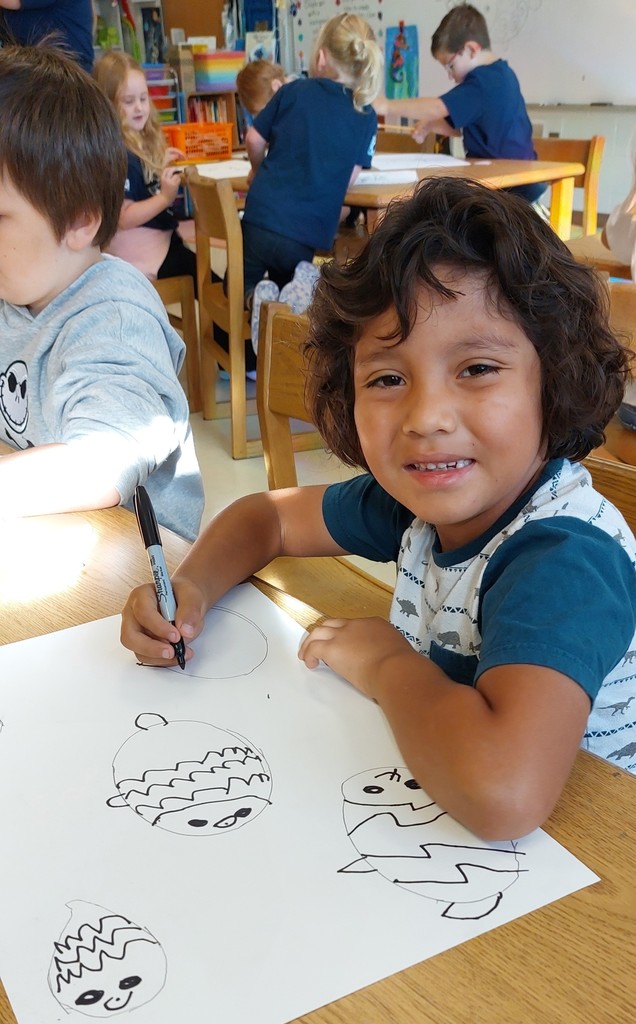 A boy is looking up.  He has a marker in his hand and his paper in front of him that he is drawing fish on.
