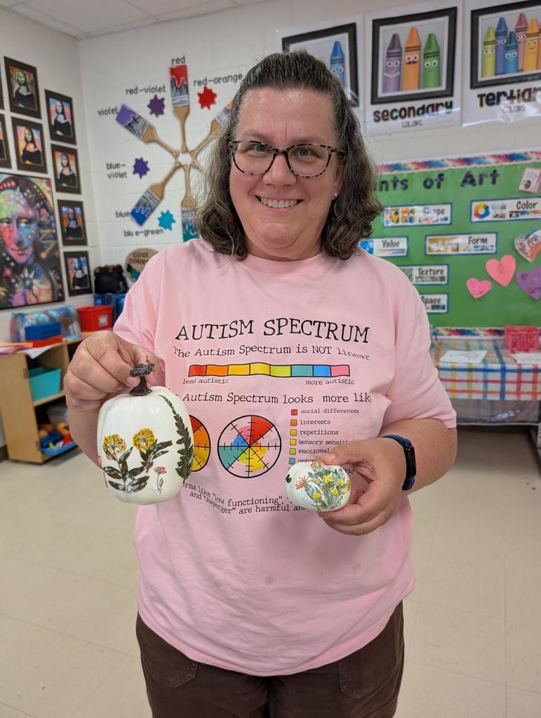 a teacher in a pink shirt holding up two pumpkins she decorated