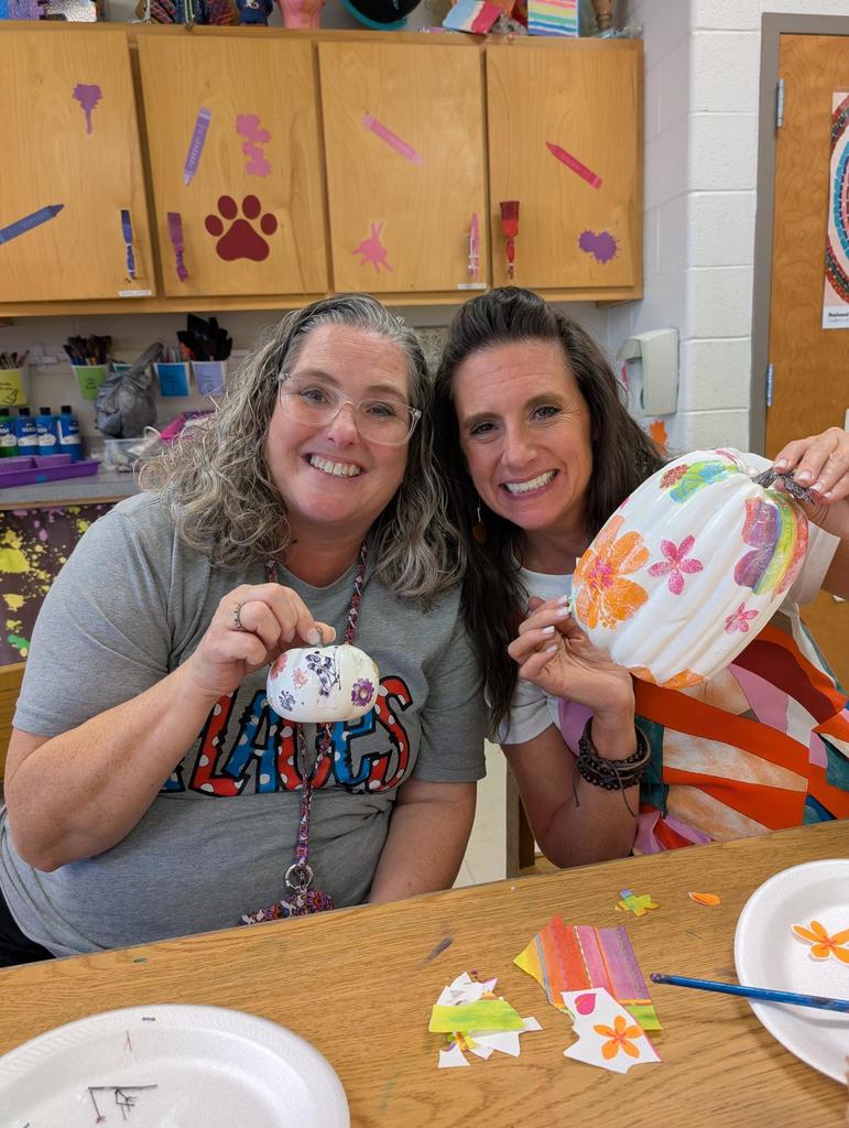 two teachers sitting at the table holding up their decorated pumpkins