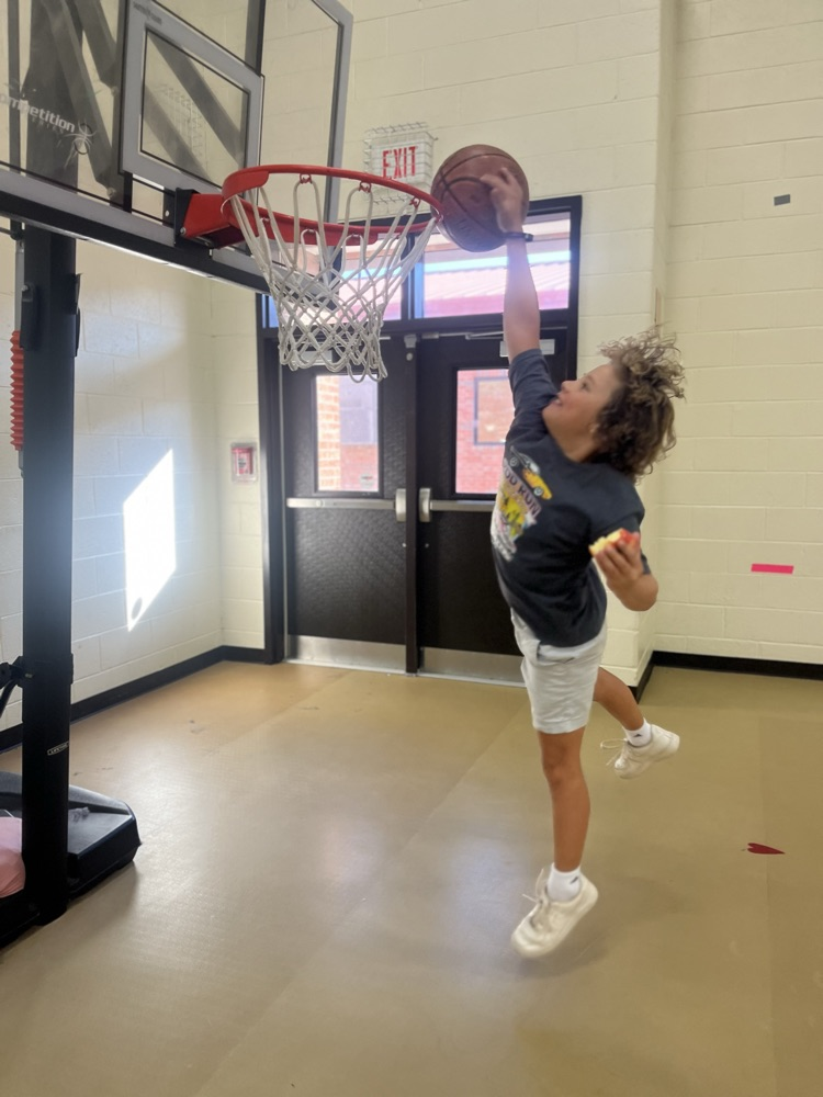 Student jumping to get basketball in goal. The student is holding an apple in one hand, his feet are off the ground, and the location is an indoor gym.