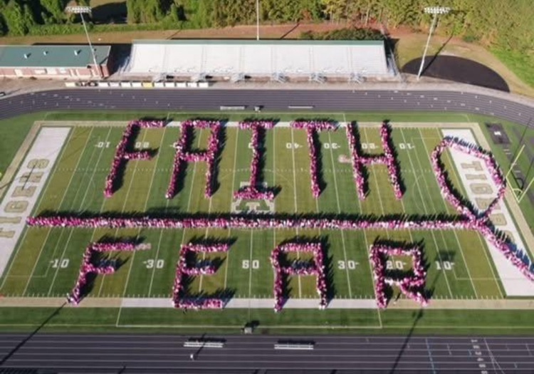 This aerial photo captures a large group of people standing on a football field arranged to form the words faith and fear with a horizontal line that separates the words that ends in a pink breast cancer ribbon so that the message reads faith over fe