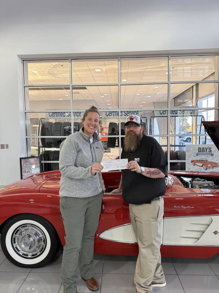 2 people standing infront of an old classic car that is red and white, the people are holding a check 