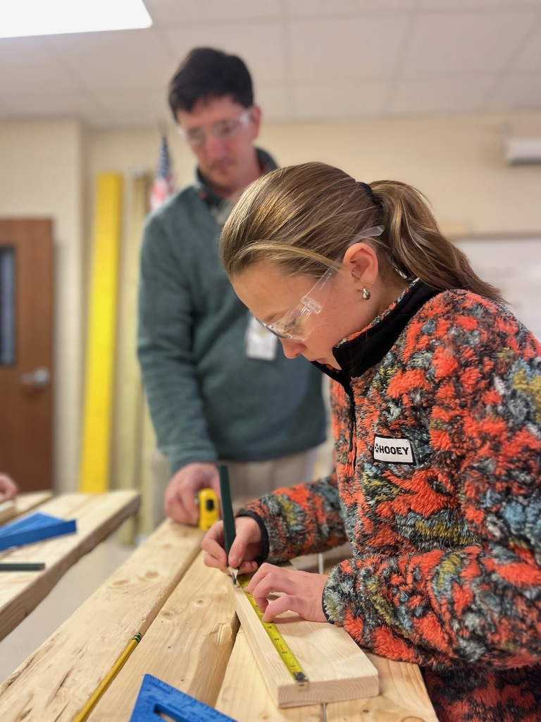 Student in foreground measures board while teacher in background assists with measuring tape.