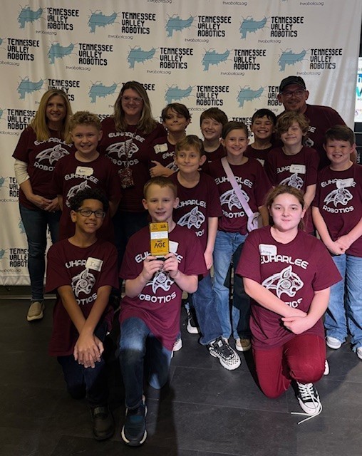 There are a group of eleven students with two teachers and a parent volunteer.  They are all wearing shirts that say, "Euharlee Robotics," on them with a wildcat in the middle.  Behind them is a backdrop that says Tennessee Valley Robotics.