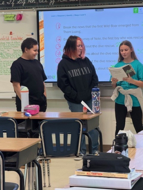 Two girls and one boy are standing in front of a classroom talking.