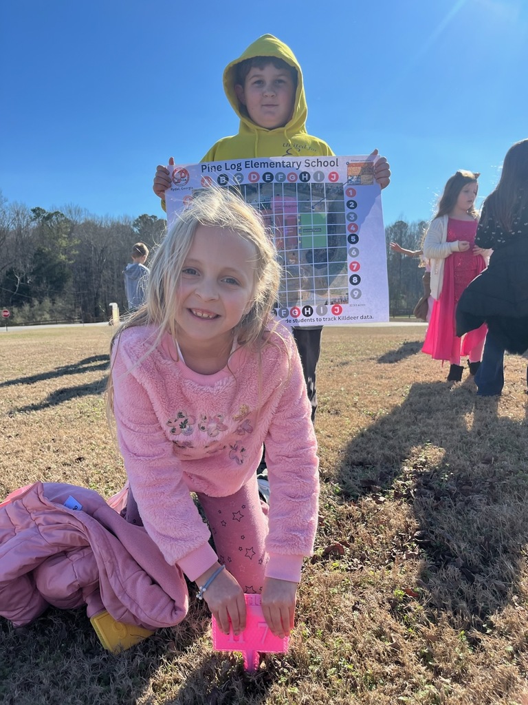 Pine Log Elementary students participate in an outdoor activity. One student kneels in the grass with a small pink plastic grid marker, while another stands behind holding a colorful Pine Log Elementary School map with grid overlay of the school. Several other children are visible in the background enjoying the sunny day
