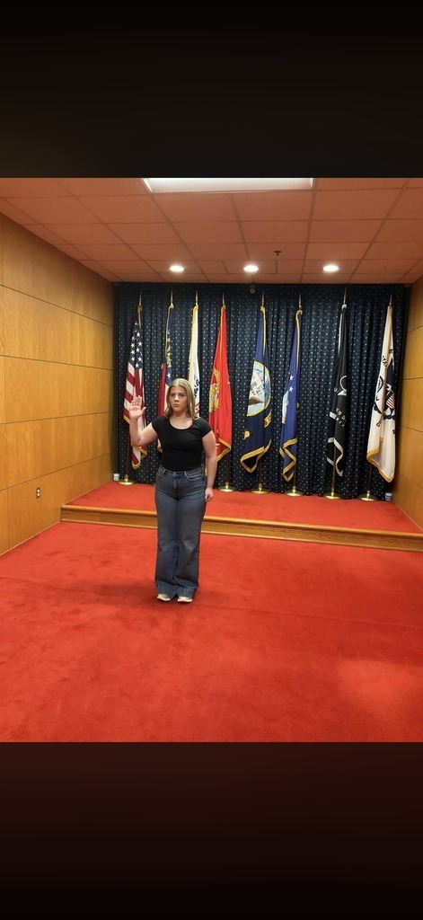 A woman stands in a room with a red floor, wearing a black shirt and jeans. Behind her, flags of various countries are displayed.
