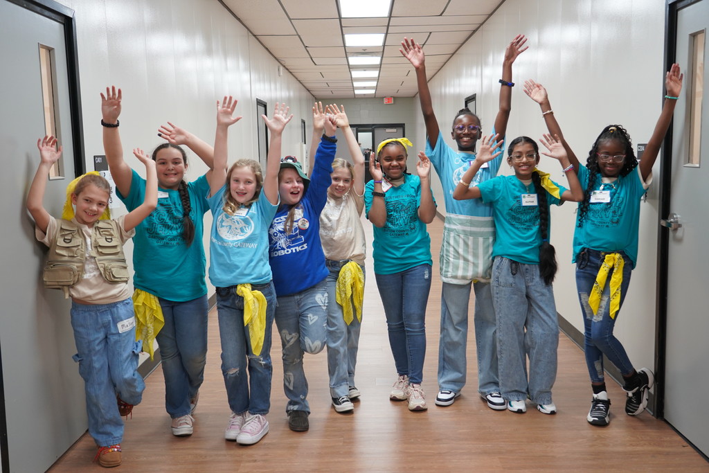 Group of young girls in matching teal shirts, posing with raised arms in a hallway.