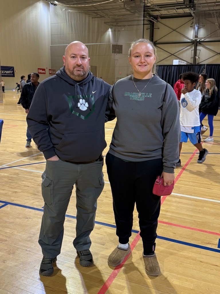 A man and a woman stand together in an indoor basketball court with blue lines, wearing gray hoodies.