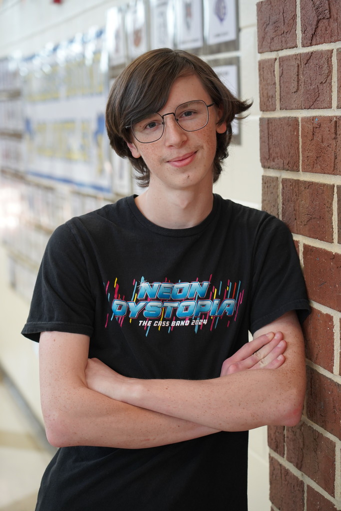 Teen with glasses and a black shirt featuring "Neon Dystopia" poses with crossed arms against a brick wall.