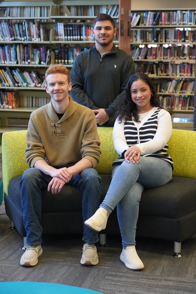 Three people sit on a couch in a library. Two are on the couch and one stands behind them.
