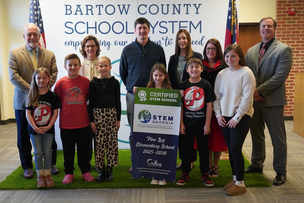 A group of people stands in front of a banner, holding a certificate. They include children, adults, and educators.