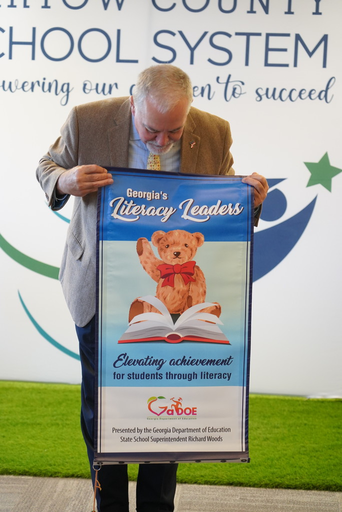 A man in a suit holds a sign featuring a bear reading a book, set against a backdrop with a green background and a banner.