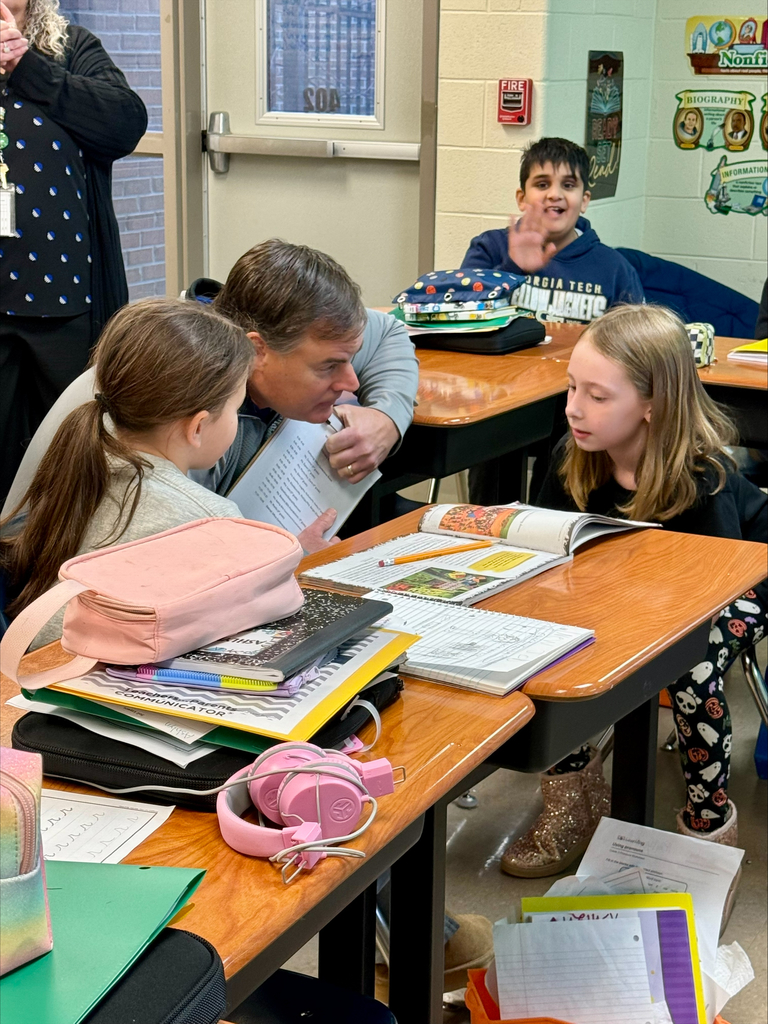 Two students sit at desks, one looking at a book, another with a backpack. An adult bends over a table, looking at the book.
