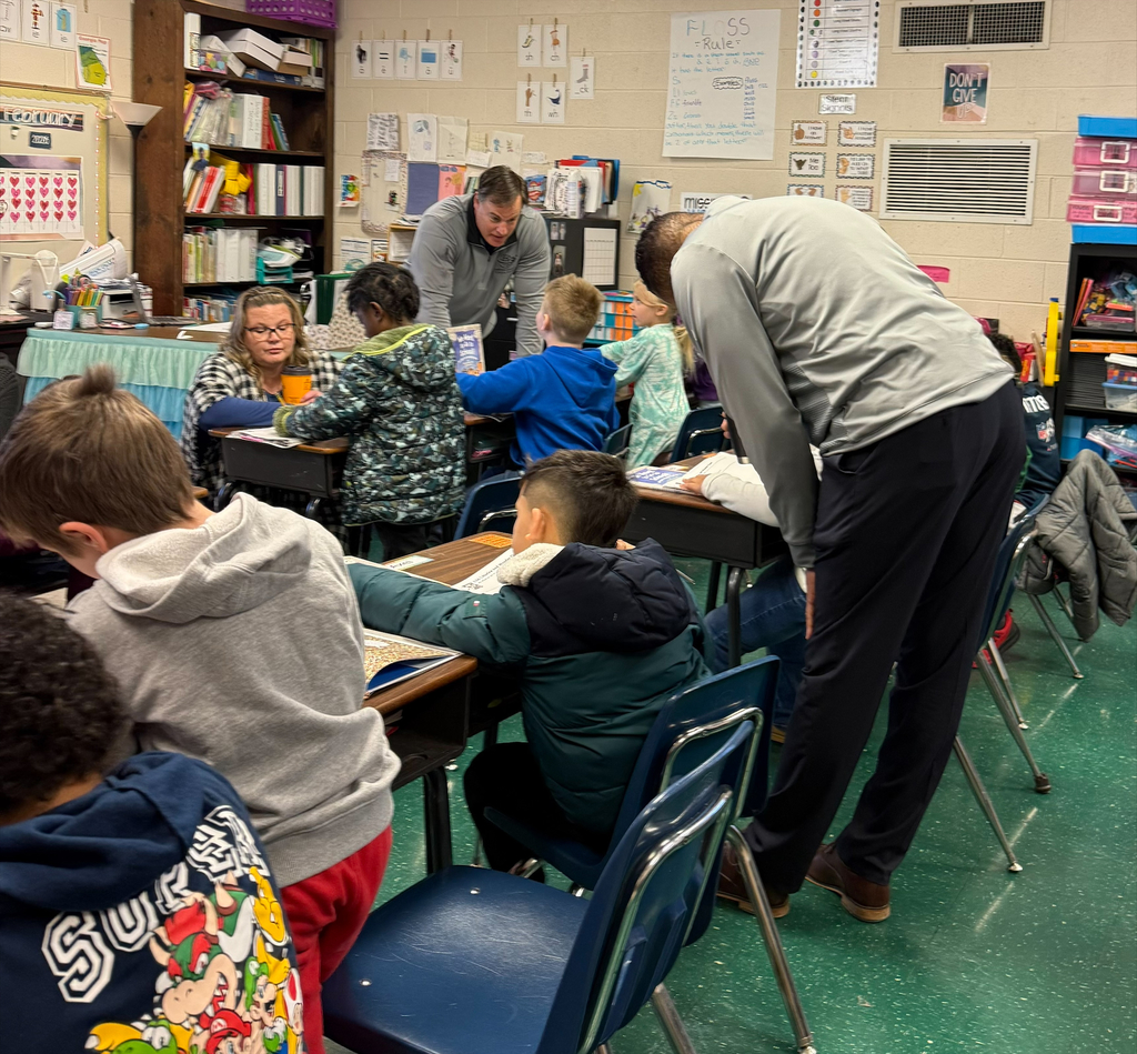 Classroom with students seated at desks, two adults helping, one leaning over a desk, shelves and books in background.
