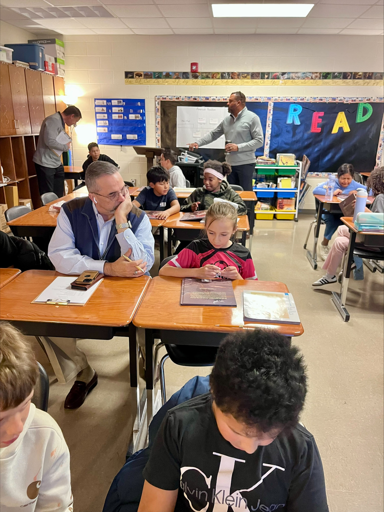 Classroom with desks and chairs, teacher stands at front, students engaged in reading or writing.