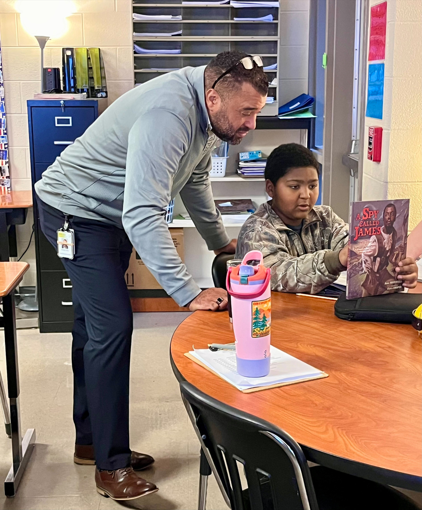 Man in gray jacket and sunglasses helps a boy with a book at a table with a lamp.