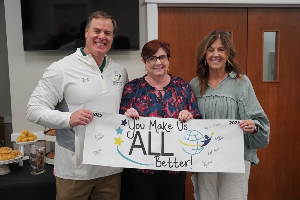 Three people holding a sign that says, "You Make Us ALL Better!" in front of a wooden door.