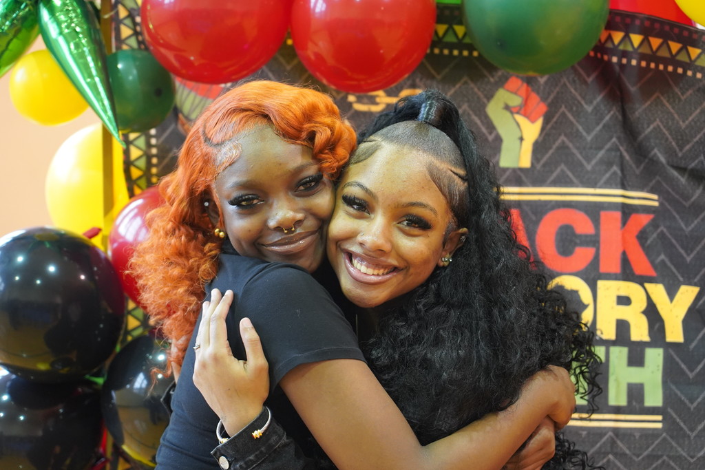 Two women hugging, smiling, and posing in front of balloons and a banner reading "Black History."
