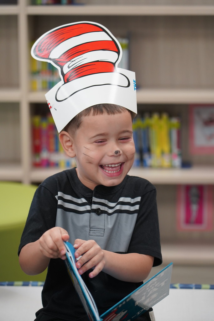 A child wears a Cat in the Hat paper hat while reading a book. Shelves with books in the background.