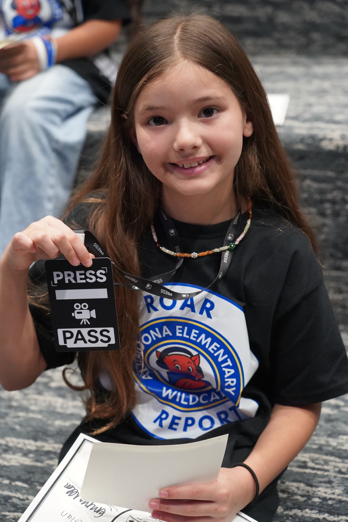 Girl with long dark hair holds a black "Press Pass" badge and paper in both hands.