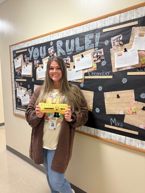 Woman wearing brown shirt and blue pants is holding a yellow ruler stands in front of a bulletin board with text and photos.