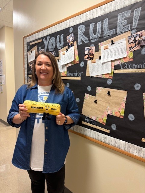 Woman in blue shirt and black pants is holding a yellow ruler stands in front of a bulletin board with text and photos.