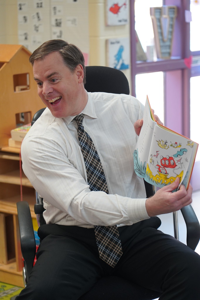 A man wearing a white shirt and tie sits in a chair, holding an open book with colorful pictures.