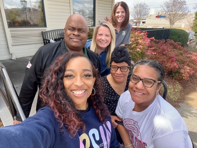 Five people stand outside a building, smiling. Two women wear glasses, one has curly hair, and two wear white shirts.