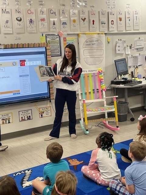 High school cheerleader reading a book to a kindergarten classroom.