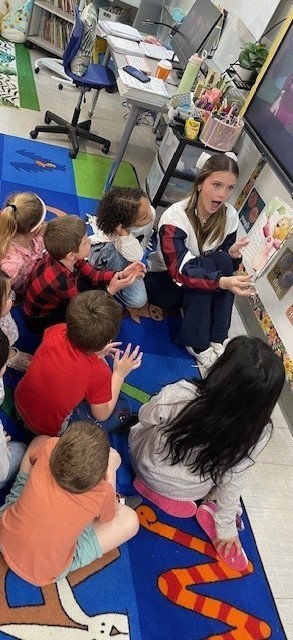 High school cheerleader reading to a group of first grade students.