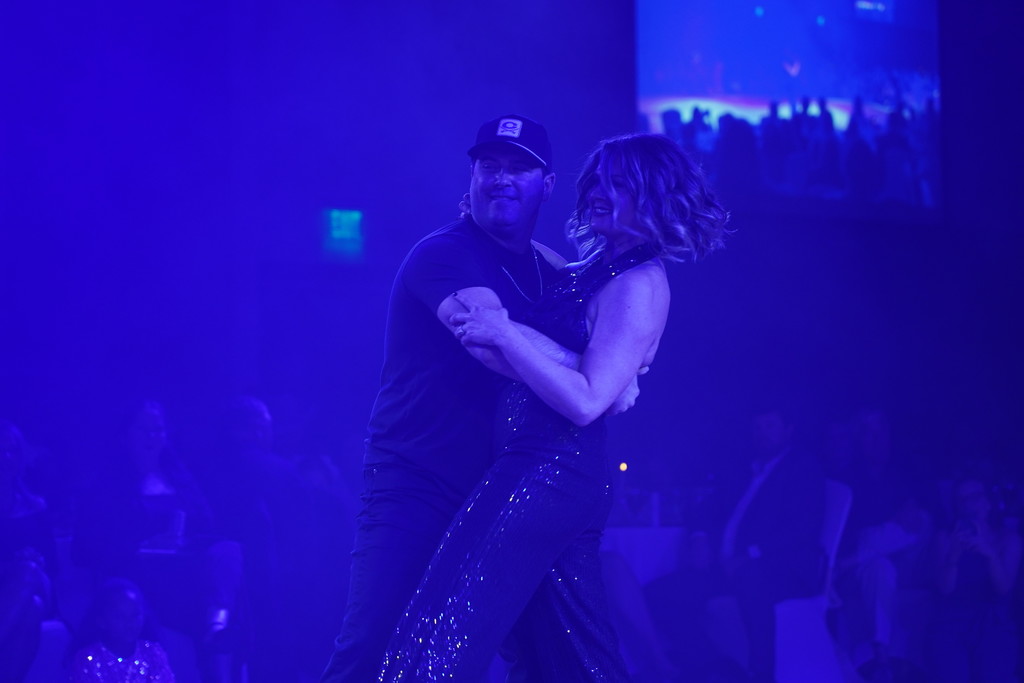 A man and woman dance on a stage under blue lighting. Audience members sit in the background.