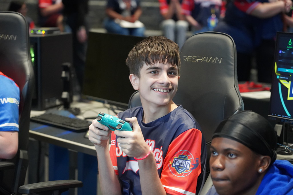 Two boys in front of computers, one wearing a red, white, and blue jersey, smiling, holding a game controller.